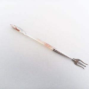 May include: A vintage cocktail fork with a clear handle and a silver-toned metal fork end. The handle has a pinkish hue near the tip and a decorative silver band. The fork has three tines.