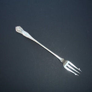 May include: A silver fork with a decorative handle and three tines. The handle has an ornate floral design and a textured, elongated stem. The fork is set against a dark background.
