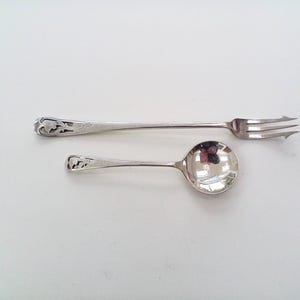 May include: A silver-toned metal fork and a matching spoon, both featuring intricate openwork scroll designs on their handles. The fork has three tines, and the spoon has a round bowl. Both pieces exhibit a polished, reflective finish.