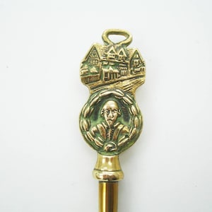 Könnte beinhalten: Ein Vintage-Brieföffner aus Messing mit einem detaillierten Relief eines Gebäudes und einem Porträt eines Mannes in einem Lorbeerkranz. Der Öffner hat oben eine Schlaufe und einen langen, schlanken Griff.