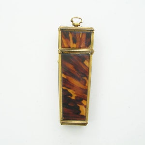 Könnte beinhalten: Ein rechteckiges antikes Etui mit Schildpattmuster und goldfarbenem Besatz. Das Etui hat oben eine kleine Schlaufe und scheint angelenkt zu sein. Das Schildpatt-Design weist Braun- und Beigetöne auf.