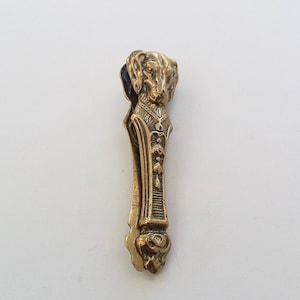 May include: A vintage gold-toned metal clip featuring a detailed dog head design at the top. The clip has an ornate, textured body with decorative patterns and a floral-like design at the base. The clip is likely used for holding papers or other small items.