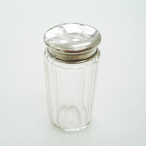 May include: A clear glass jar with a silver lid. The jar has vertical ridges and a silver band around the top. The lid is circular with a patterned design. The jar is likely for storing cosmetics or other small items.