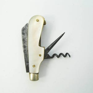 May include: A vintage wine opener with a cream-colored handle and brass accents. The opener features a blade, a corkscrew, and a small metal piece. The tool is designed for opening wine bottles.