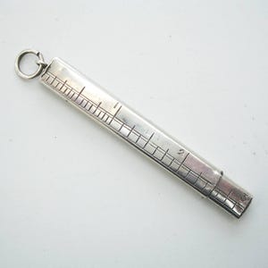 Könnte beinhalten: Ein silberner Taschenlineal mit einem Ring oben. Das Lineal ist rechteckig mit geätzten Markierungen und Zahlen. Das Lineal misst bis zu 5,08 cm.