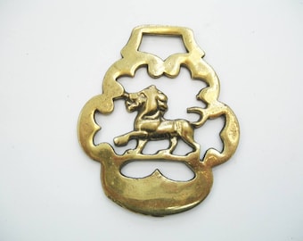 Puede incluir: Un herraje de latón antiguo para caballo con un león. El león está representado dentro de un marco calado ornamental. El latón tiene un tono dorado y muestra signos de edad, lo que sugiere su origen vintage. El diseño es simétrico.