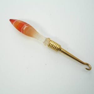 Könnte beinhalten: Ein Häkelhaken mit dekorativem Griff. Der Griff hat orange und weiße Streifen. Der Haken besteht aus goldfarbenem Metall.