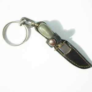 Può includere: Un portachiavi decorativo con un coltello in miniatura. Il coltello ha una lama argentata e un fodero in pelle nera con cuciture dorate. L'impugnatura è verde chiaro e l'anello portachiavi è argentato.