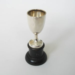May include: A small silver cup with a rounded bowl and a slender stem, mounted on a dark, circular wooden base. The cup has a polished, reflective surface, and the base is a deep, contrasting black.