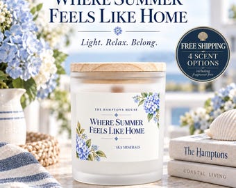May include: A frosted glass candle jar with a light wood lid. The label features blue hydrangea illustrations and the text: THE HAMPTONS HOUSE, WHERE SUMMER FEELS LIKE HOME, and SEA MINERALS. The candle is positioned near books and blue flowers.