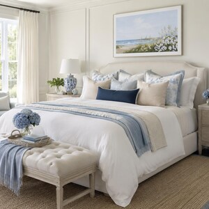 May include: A bedroom featuring a large bed with white bedding, layered with blue and beige throw blankets and pillows. A tufted beige bench sits at the foot of the bed. Two blue and white patterned lamps rest on wooden nightstands.