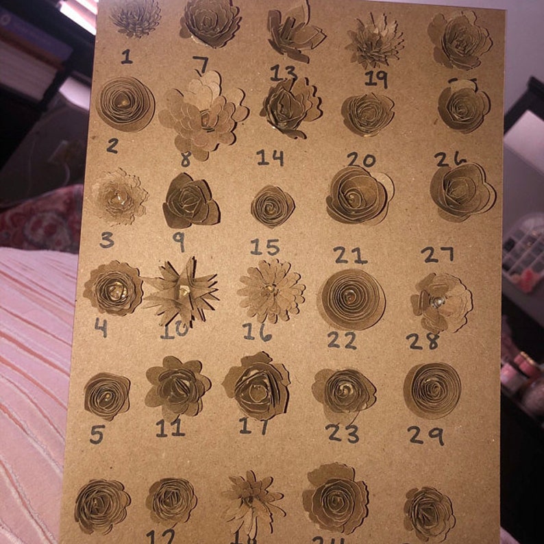 Op de afbeelding: Een bruin papieren ambacht met 29 genummerde, handgemaakte papieren bloemen in diverse ontwerpen. De bloemen zijn gerangschikt in rijen en kolommen op een kartonnen oppervlak, met een verscheidenheid aan vormen en texturen.