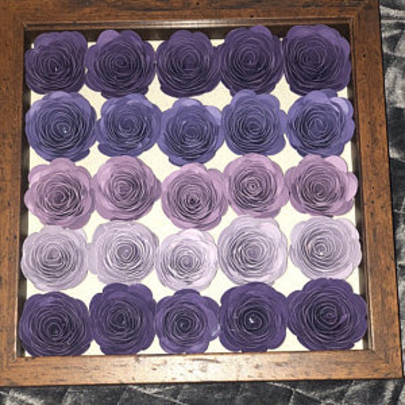Op de afbeelding: Een ingelijst kunstwerk met een raster van papieren rozen in verschillende tinten paars, van diep violet tot licht lavendel. De rozen zijn in rijen gerangschikt in een vierkante, houten lijst.