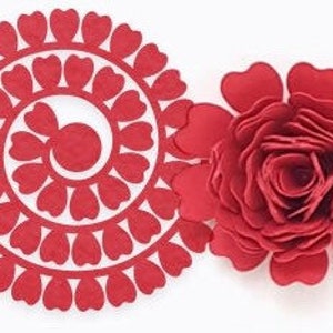 Op de afbeelding: Twee rode papieren bloemdecoraties. E&eacute;n is een gelaagde roos en de andere is een cirkelvormig ontwerp met hartvormige uitsparingen. Beide staan op een witte achtergrond. De roos heeft meerdere lagen bloemblaadjes.