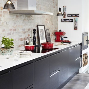 K&ouml;nnte beinhalten: Eine moderne K&uuml;che mit schwarzen Schr&auml;nken, einer wei&szlig;en Arbeitsplatte und einer Backstein-R&uuml;ckwand. Auf dem Herd stehen zwei rote T&ouml;pfe, eine Kaffeemaschine und ein gerahmtes Bild an der Wand. Der Text "Brooklyn" und "New York City" ist an der Wand zu sehen.