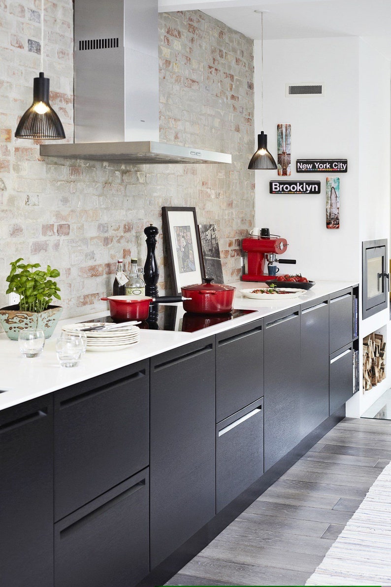 K&ouml;nnte beinhalten: Eine moderne K&uuml;che mit schwarzen Schr&auml;nken, einer wei&szlig;en Arbeitsplatte und einer Backstein-R&uuml;ckwand. Auf dem Herd stehen zwei rote T&ouml;pfe, eine Kaffeemaschine und ein gerahmtes Bild an der Wand. Der Text "Brooklyn" und "New York City" ist an der Wand zu sehen.