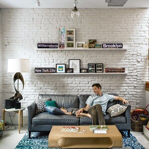K&ouml;nnte beinhalten: Ein Wohnzimmer mit einer wei&szlig;en Backsteinwand, einem grauen Ledersofa, einem Holz-Couchtisch und einem blau-wei&szlig; gemusterten Teppich. Auf dem Sofa schl&auml;ft ein Kind und ein Mann sitzt auf dem Sofa. An der Wand befinden sich Regale mit gerahmten Fotos und Schildern mit der Aufschrift "Williamsburg", "New York City", "Brooklyn" und "Nicht anlehnen".