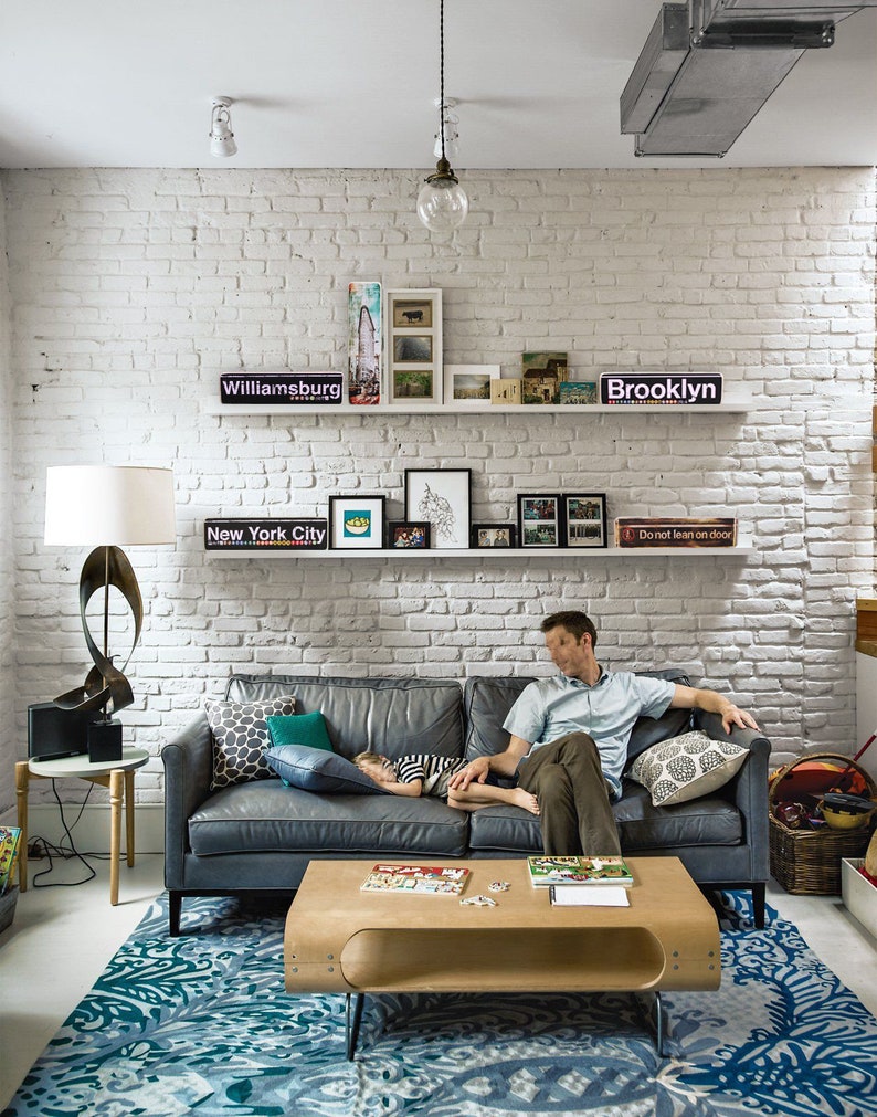 K&ouml;nnte beinhalten: Ein Wohnzimmer mit einer wei&szlig;en Backsteinwand, einem grauen Ledersofa, einem Holz-Couchtisch und einem blau-wei&szlig; gemusterten Teppich. Auf dem Sofa schl&auml;ft ein Kind und ein Mann sitzt auf dem Sofa. An der Wand befinden sich Regale mit gerahmten Fotos und Schildern mit der Aufschrift "Williamsburg", "New York City", "Brooklyn" und "Nicht anlehnen".