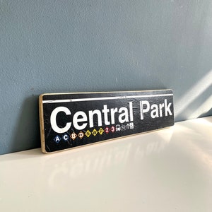 Op de afbeelding: Een rechthoekig bord met de tekst "Central Park" in witte letters op een zwarte achtergrond. Daaronder gekleurde iconen en letters. Het houten bord heeft een verweerde uitstraling.