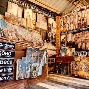 K&ouml;nnte beinhalten: Eine Wandanzeige aus Holz mit Schildern und Bildern von New Yorker Stra&szlig;en im Vintage-Stil. Die Schilder zeigen Namen von Stadtteilen wie Broadway, Soho, Tribeca, Little Italy und Fashion Avenue. Die Bilder zeigen die Brooklyn Bridge, das Empire State Building und die Freiheitsstatue.
