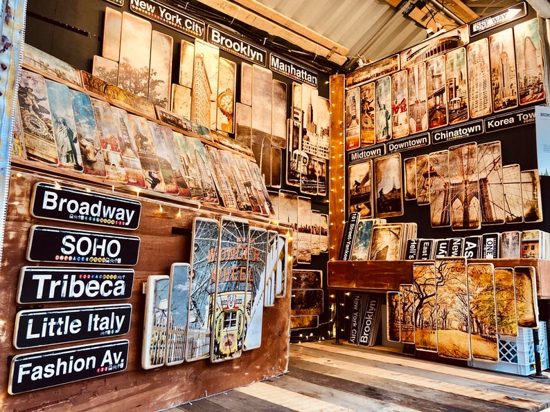 K&ouml;nnte beinhalten: Eine Wandanzeige aus Holz mit Schildern und Bildern von New Yorker Stra&szlig;en im Vintage-Stil. Die Schilder zeigen Namen von Stadtteilen wie Broadway, Soho, Tribeca, Little Italy und Fashion Avenue. Die Bilder zeigen die Brooklyn Bridge, das Empire State Building und die Freiheitsstatue.
