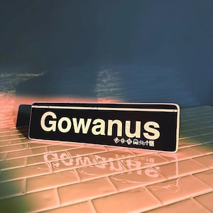 以下が含まれることがあります： 白黒の街路標識に白い文字で「Gowanus」と書かれている。標識はタイル張りの表面にある。