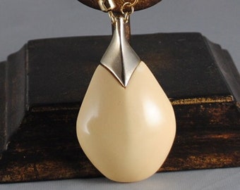 May include: A gold chain necklace with a teardrop-shaped pendant. The pendant is made of a cream-colored material and has a silver-toned top.