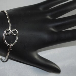 May include: A silver bangle bracelet with a heart-shaped charm. The charm is made of two interlocking circles.
