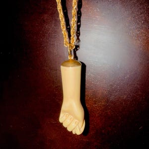 May include: A gold-colored chain necklace with a pendant shaped like a clenched fist. The pendant is a light beige color, with a gold-colored cap at the top. The chain has a rope-like design.
