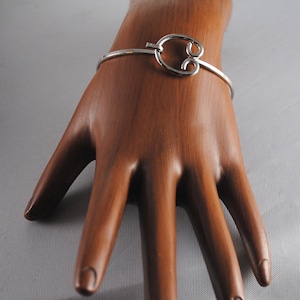 May include: A silver bangle bracelet with a unique, open, figure-eight design. The bracelet is shown on a wooden hand model.