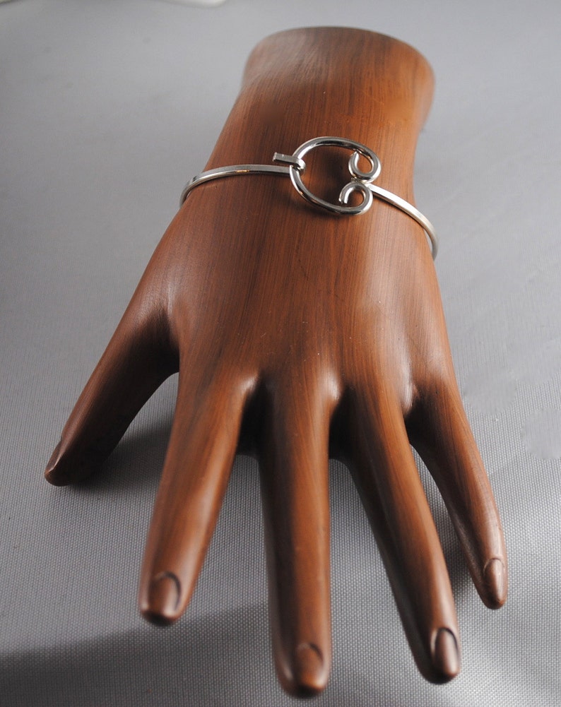 May include: A silver bangle bracelet with a unique, open, figure-eight design. The bracelet is shown on a wooden hand model.