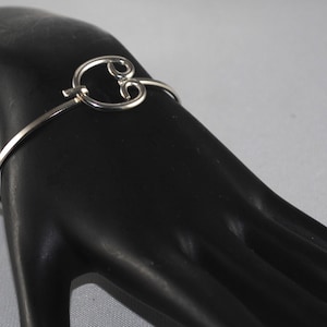 May include: A silver bangle bracelet with a decorative, open circle design. The circle is made of two overlapping loops.