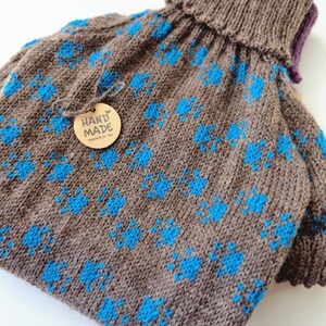 May include: A handmade brown knitted hot water bottle cover with a blue floral pattern. The cover has a ribbed turtleneck collar and a small tag that says "HAND MADE Especially for You".