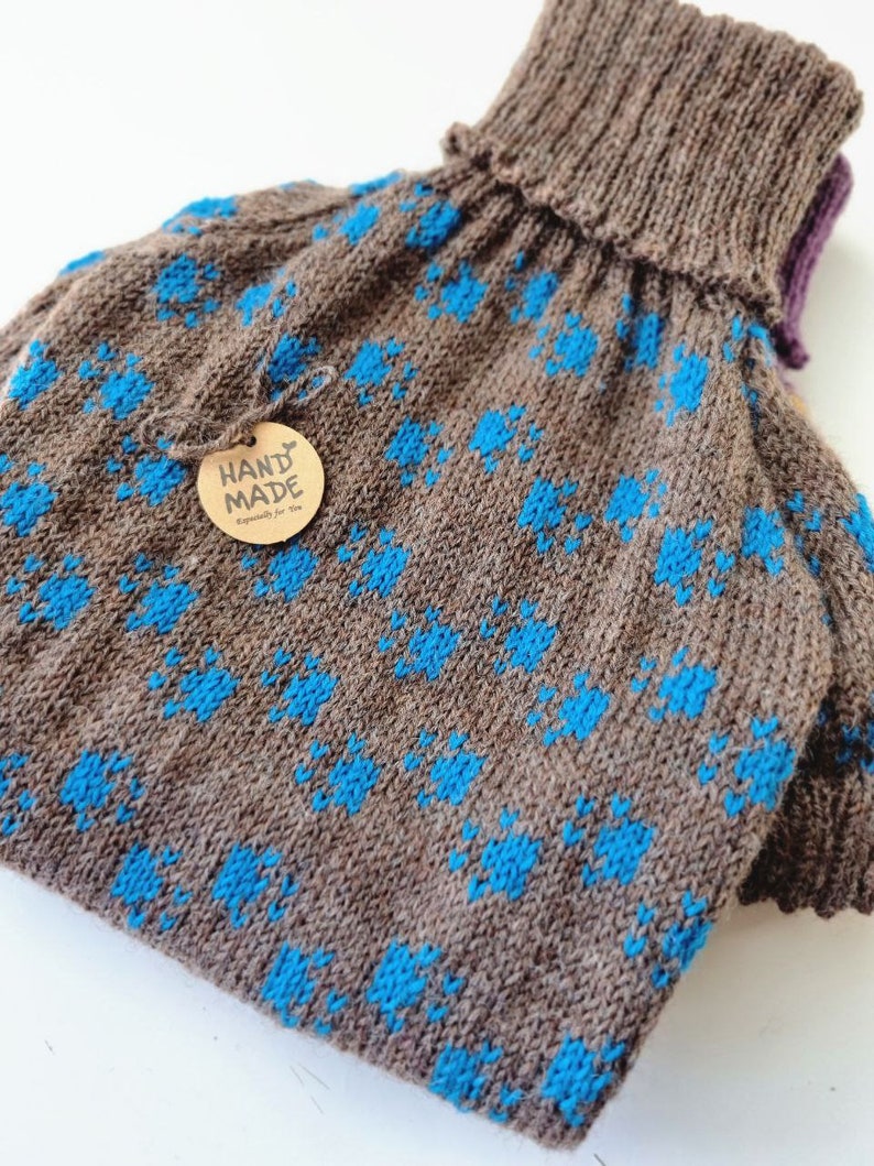 May include: A handmade brown knitted hot water bottle cover with a blue floral pattern. The cover has a ribbed turtleneck collar and a small tag that says "HAND MADE Especially for You".