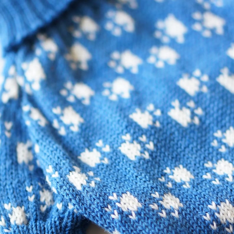 May include: Close-up of a blue knitted fabric with a repeating white paw print pattern. The knitwear has a textured appearance, with the white paw prints evenly spaced across the blue background. The fabric appears soft and cozy.