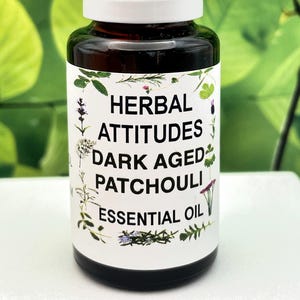 May include: A dark amber glass bottle with a white cap, labeled "Herbal Attitudes Dark Aged Patchouli Essential Oil." The label is decorated with botanical illustrations. The bottle is filled with a dark liquid.