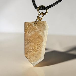 May include: A pendant necklace featuring a light brown, rectangular stone with a unique pattern. The stone is suspended from a gold-colored ring and chain, attached to a black cord. The pendant has a pointed bottom.