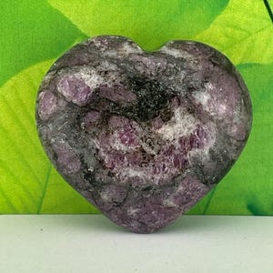 May include: A heart-shaped stone with a mix of dark gray, white, and deep pink hues. The stone has a textured surface and a natural, organic appearance. The heart shape is clearly defined, making it a unique decorative item.