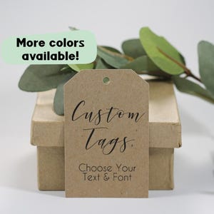 May include: A brown kraft paper tag with the words "Custom Tags" in elegant script and "Choose Your Text & Font" below. The tag is on a small cardboard box, with a sprig of greenery in the background. A green oval says "More colours available!"
