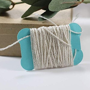May include: A teal blue card holds a spool of natural twine. The twine is a light beige colour and is neatly wound. A small brown box is in the background, with green leaves in the upper left corner. Ideal for crafting and gift wrapping.