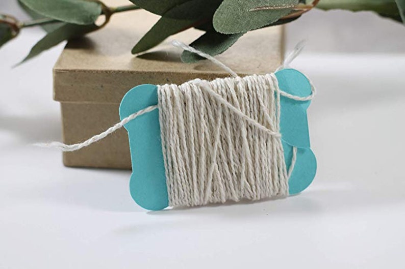 May include: A teal blue card holds a spool of natural twine. The twine is a light beige colour and is neatly wound. A small brown box is in the background, with green leaves in the upper left corner. Ideal for crafting and gift wrapping.