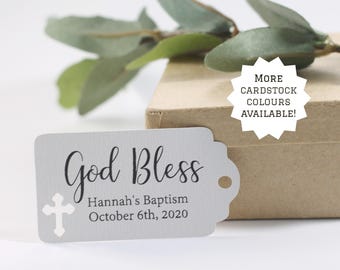 May include: A light gray baptism tag with the words "God Bless" in elegant script, along with "Hannah's Baptism October 6th, 2020." A white cross is on the left. The tag is near a brown box and green leaves.