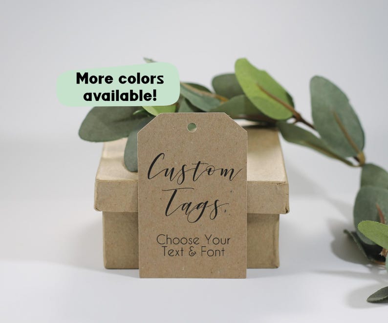 May include: A brown kraft paper tag with the words "Custom Tags" in elegant script and "Choose Your Text & Font" below. The tag is on a small cardboard box, with a sprig of greenery in the background. A green oval says "More colours available!"