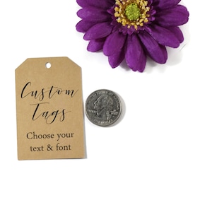 May include: A brown custom tag with the text "Custom Tags" and "Choose your text & font" in black script. A purple flower and a quarter coin are also in the image.