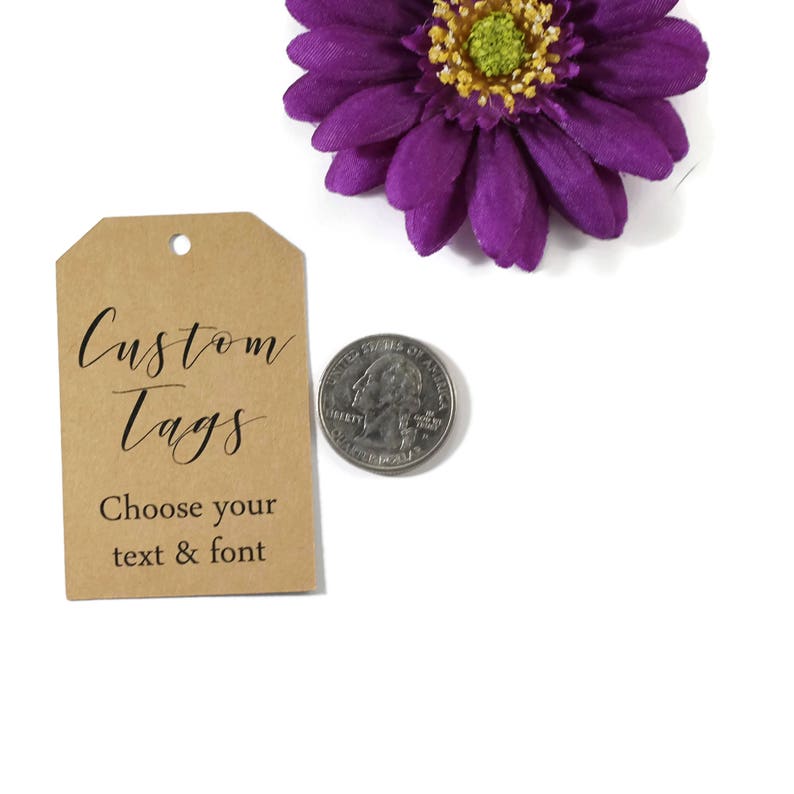 May include: A brown custom tag with the text "Custom Tags" and "Choose your text & font" in black script. A purple flower and a quarter coin are also in the image.