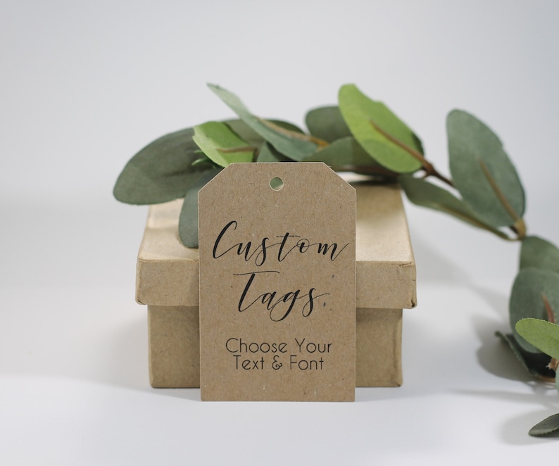 May include: A brown kraft paper tag with the words "Custom Tags" in elegant black script. Below, the text reads "Choose Your Text & Font." The tag is positioned in front of a small, light brown box, with green leaves in the background.