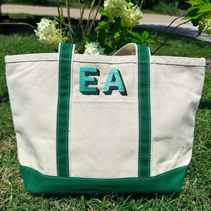 May include: A canvas tote bag with a cream-colored body, green accents, and handles. The bag features the embroidered letters "EA" in a teal and black 3D design. The bottom and trim are green.
