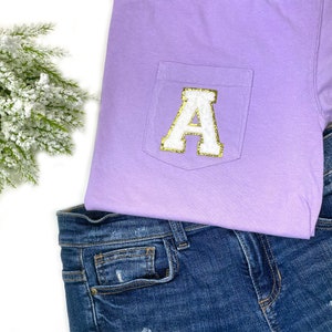 May include: A lavender t-shirt with a white pocket featuring a gold glitter letter A.