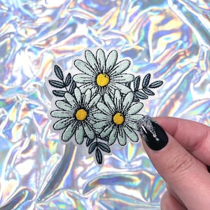 May include: Embroidered patch featuring three daisies with light blue petals and yellow centers, surrounded by green leaves. The patch is white with black outlines and is held against a shimmering, iridescent background.