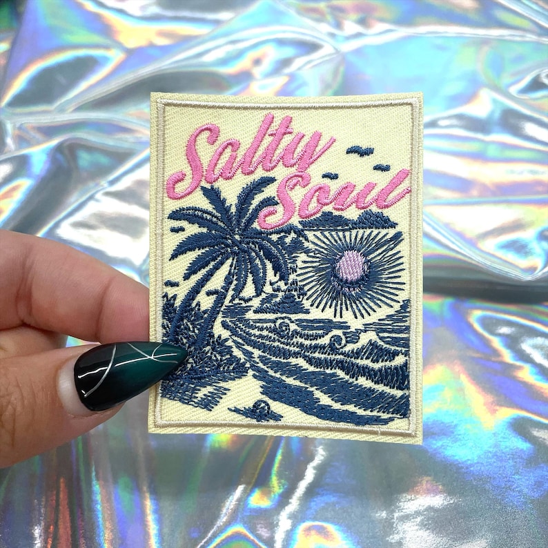 May include: Embroidered patch with the words "Salty Soul" in pink script above a blue and white beach scene with a palm tree, waves, and a sun. The patch has a beige background and a rectangular shape.